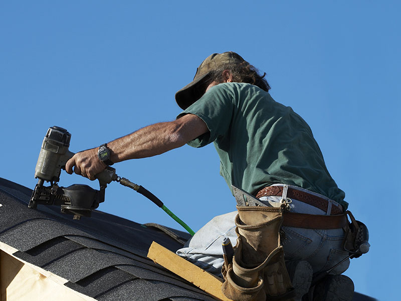 Close-up of a worker installing dark gray shingles on a residential roof using a nail gun